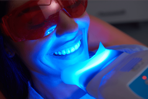 A patient wearing protective red goggles receiving a blue light teeth whitening treatment in a dental office.