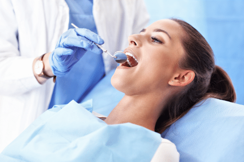 A dental professional performs a procedure on a reclining patient in a clinical setting, using instruments while wearing a white coat, blue scrubs, and gloves.