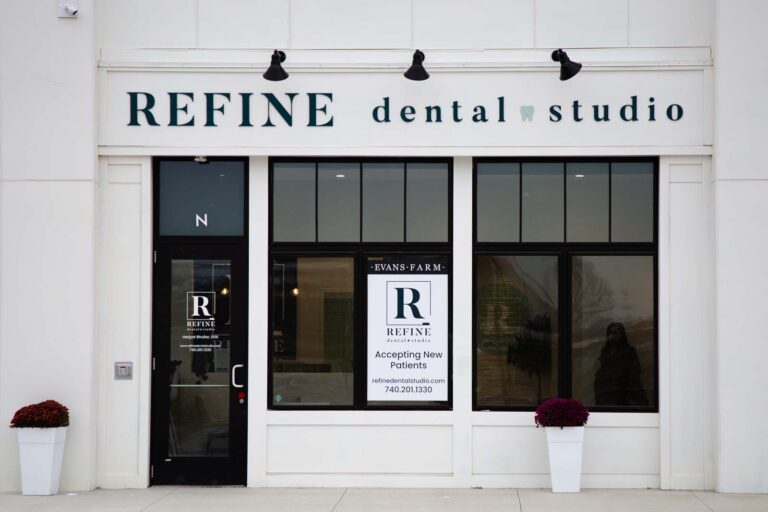 The storefront of Refine Dental Studio features large windows and a glass door, with a modern and inviting design. Signs display the clinic's name, logo, dentist's information, and contact details.