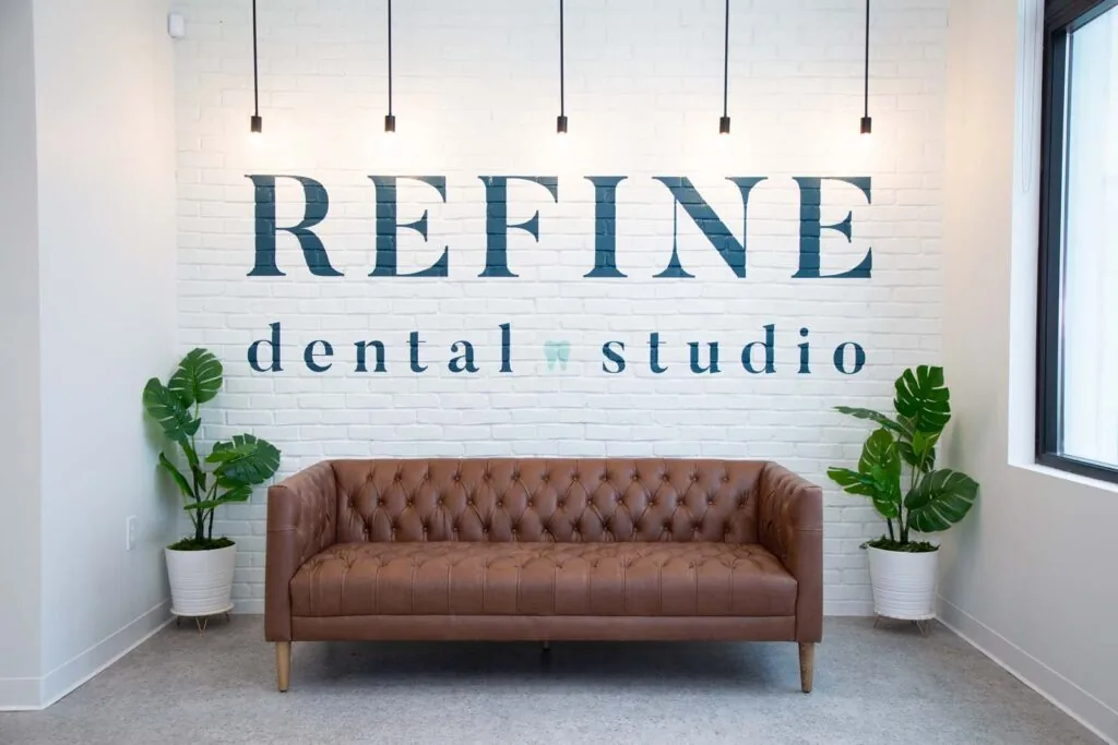 A modern dental studio waiting area featuring a brown tufted leather sofa against a white brick wall. The wall displays the logo "REFINE dental studio" with a tooth icon, accompanied by two potted plants and pendant lighting for a welcoming atmosphere.